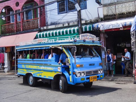 Songthaew azul y amarillo con pasajeros sentados en bancos de madera, en una calle de Phuket Town